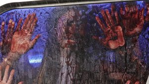 Plan issu du film Massacre à la tronçonneuse (2022) : de l'intérieur une femme a le visage et les paumes des mains collés contre la vitre d'une voiture, du sang coule sur le verre de partout.