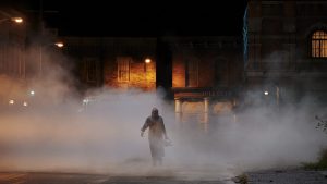 La silhouette de Leatherface, de dos, tenant sa tronçonneuse, se dessine dans une rue embrumée, éclairée par des lampadaires jaunes, dans le film Massacre à la tronçonneuse (2022).