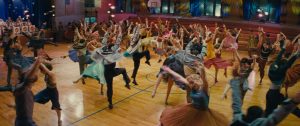Scène de danse collective sur un terrain de basket, où tous les danseurs, hommes et femmes, portent des tenues de bal colorées ; scène du film West Side Story.
