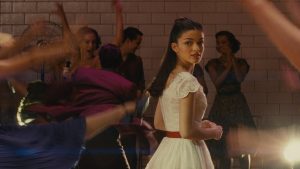 En plein bal, au milieu d'individus flous, la silhouette nette de Maria, qui se tourne vers nous, avec sa robe blanche ; plan issu du film West Side Story 2021.