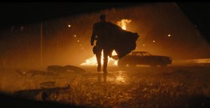 La silhouette de The Batman se dessine, s'approchant vers nous, la cape au vent ; derrière lui, une voiture prend feu dans une rue inondée d'une lumière jaune.