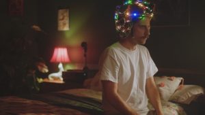 Un homme est assis sur le bord de son lit, les yeux clos, portant une guirlande allumée tout autour du crâne, dans le film Strawberry Mansion.