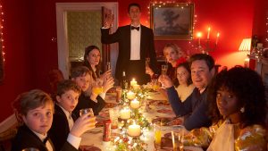 Tout autour de la longue table, les dix convives de la soirée de Noël du film Silent Night nous regardent, interloqués, ; en bout de table, un homme en costume noeud papillon porte un toast ; la scène se situe dans un salon aux murs rouges et avec de nombreuses bougies à la lueur jaune.