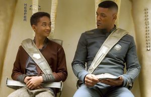 Jada et Will Smith assis côte à côté, en combinaison de science-fiction, dans le film After Earth ; le père a un livre dans ses mains, il fait la leçon à son fils qui le regarde avec malice.