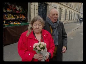 Françoise Lebrun et Dario Argento dans les rues de Paris, Lebrun tient un bouquet de fleurs, Argento suit inquiet derrière ; scène du film Vortex Gaspar Noé.