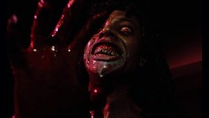 Plongé dans un violent clair-obscur noir et rouge, une femme démon ensanglantée et au visage possédé regarde droit dans l'objectif de la caméra du film Démons.