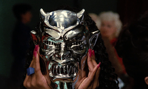 Plan issu du film Démons où une femme se cache derrière un masque de démon en argent.