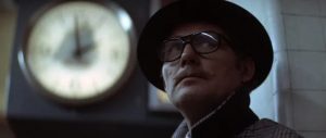 Plan rapproché-épaule sur le visage de Robert Shaw, concentré, portant un chapeau, une moustache et des lunettes ; à l'arrière-plan l'horloge d'une station de métro ; plan du film Les pirates du métro.