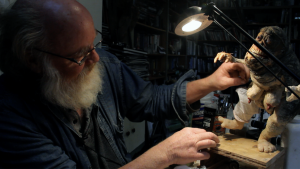 Phil Tippett travaille à son bureau sur une maquette de monstre lézard avec des seins féminins proéminents, à la lueur d'une petite lampe de bureau ; scène de tournage du film Mad god.
