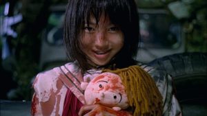 Une petite fille affiche un sourire large sourire enfantin, alors que son visage et la poupée qu'elle tient dans ses mains sont tachés de sang dans le film Battle Royale.