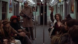 Dans une rame de métro plusieurs passagers assis et inquiets sont surveillés par un homme portant des lunettes de soleil noires, un long manteau, et une mitraillette ; scène du film Les pirates du métro (1974).