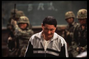 Au premier plan, Takeshi Kitano, le visage narquois incliné vers le sol ; derrière lui un groupe de militaires devant un tableau d'écolier vert ; scène du film Battle Royale.