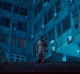 En contre-plongée, un homme, la tête baissée, vêtu d'un uniforme gris-jaune, s'apprête à descendre un escalier ; derrière lui des immeubles vétustes, baignés d'une lumière bleue futuriste ; scène du film Underdog.