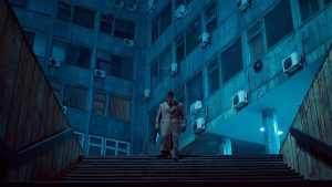 En contre-plongée, un homme, la tête baissée, vêtu d'un uniforme gris-jaune, s'apprête à descendre un escalier ; derrière lui des immeubles vétustes, baignés d'une lumière bleue futuriste ; scène du film Underdogs.