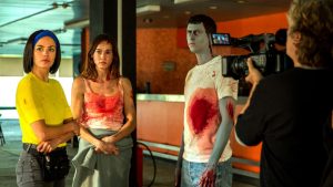 Trois comédiens dont un déguisé et maquillé en zombie attendent les bras ballants, sous l'oeil du cadreur sur le tournage du film Coupez !