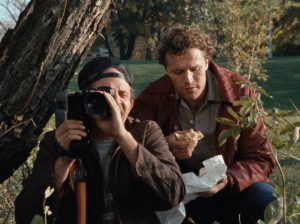 Dans un parc, tandis que son camarade filme, l'oeil bien placé dans le capteur d'un caméscope posé sur pied, Henry mange un hamburger, accroupi derrière lui, distrait ; scène du film Henry portrait d'un serial killer.