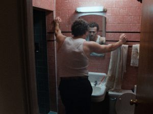Michael Rooker, vu de dos, se regarde dans le miroir, dans une salle de bains en briques rouges, dans le film Henry portrait d'un serial killer.