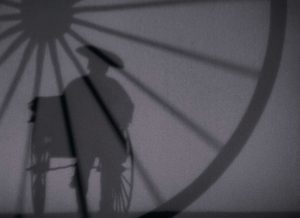 Vu en ombre chinoise, une silhouette masculine conduit un pousse-pousse ; en surimpression, une roue de pousse-pousse ; plan issu du film L'homme au pousse-pousse.
