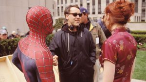 Sam Raimi sur le tournage de Spiderman, donne ses consignes à Tobey Maguire déguisé en Spiderman et Kirsten Dunst, en robe chinoise ; tous deux vus de dos.
