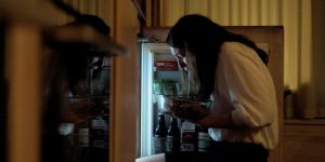 Une jeune femme brune vomit dans un saladier, devant la porte ouverte de son frigo plein ; scène de nuit du film The Feast.