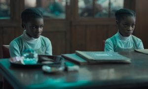 Deux petites filles afro-américaines sont assises côte à côte derrière une table en bois sur laquelle traînent deux cahiers noirs ; elles ont le visage des enfants que l'on s'apprête à disputer ; scène du film The Silent Twins.