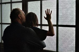 Un homme et une femme enlacés contre une large fenêtre, aux vitres opaques ; l'homme est derrière la femme qui pose sa main grande ouverte sur l'une des vitres ; toute la scène est baignée dans la faible lumière du film Inexorable.