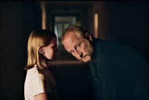 Dans un couloir sombre et indéfini, une petite fille blonde chuchote à l'oreille de Benoît Poelvoorde, penché vers elle, le visage tourné vers sa gauche ; scène du film Inexorable.