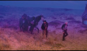 Deux femmes à l'allure de cow-boy marchent dans une plaine et tirent un cheval derrière elles ; le paysage est baignée dans une lumière violette et jaune surréaliste ; scène du film After Blue (Paradis Sale) de Betrand Mandico.