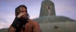 Au premier plan à gauche, Arnold Schwarzenegger, assis, regarde l'horizon, cadré en rapproché-épaule ; derrière lui, au sommet d'une colline, ce qui semble être une stèle sur laquelle est peinte une femme ; plan issu du film Conan le barbare, cité par Bertrand Mandico.