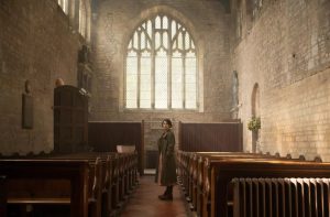 Jessie Buckley entre les rangées de sièges d'une petite église vide ; la lumière du soleil passe par les vitraux et donnent une couleur ocre au lieu ; scène du film Men.