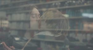 Les deux amoureux du film Mad Love in New-York des Frères Safdie s'embrassent sur la bouche ; on les voit à travers la vitre de ce qui semble être une épicerie.