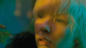 Gros plan sur le visage rêveur de Paula Luna, plongée dans une lumière évanescente jaune et vert bleuté issu du film After Blue (Paradis Sale) de Bertrand Mandico.