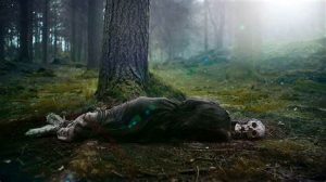 Un squelette, pieds et mains liés, pourrit au pied d'un arbre dans une forêt baigné dans une douce lumière de soleil dans le film The Green Knight.