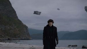 Au premier plan Timothée Chalamet la tête baissée, les bras le long du corps ; derrière lui, la plage, et dans le ciel trois vaisseaux spatiaux flottant ; scène du film Dune.