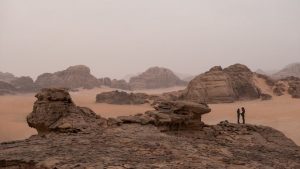 Plan d'ensemble sur un vaste désert ponctué par des roches imposantes ; en bas à droite de l'image, deux silhouettes humaines, minuscules, vues de dos ; issu du film Dune de Denis Villeneuve.