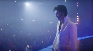 Plan rapproché-taille sur Austin Butler incarnant Elvis dans les années 70, avec la banane et sa tenue blanche, sur scène à Las Vegas ; il est debout, tourné vers le public attablé, nous le voyons de trois-quarts ; la salle est éclairée d'une lumière bleu avec des teintes rosées.