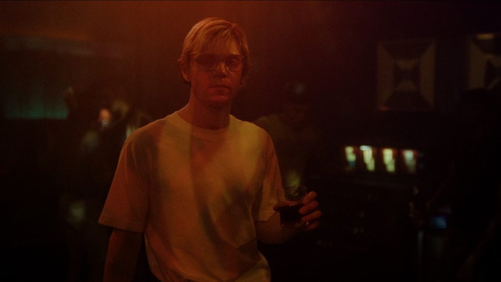 Jeffrey Dahmer en boîte de nuit, un verre d'alcool dans la main et portant un t-shirt blanc, dans une lumière rouge sombre.
