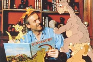 Don Bluth lit un livre sur les dinosaures, entourés des dinosaures en dessin animé qui lui grimpent dessus.