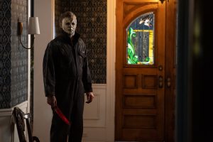 Micahel Myers se tient debout, un couteau ensanglanté dans sa main, dans le vestibule d'une maison calme, juste à côté de la porte d'entrée, comme dans l'attente ; scène du film Halloween Ends.