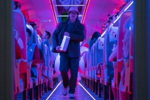 Brad Pitt marche les rangées d'un train étrangement éclairé d'une lumière rose fluo ; il porte un chapeau, des lunettes, et une valise sous le bras ; plan issu du film Bullet Train.