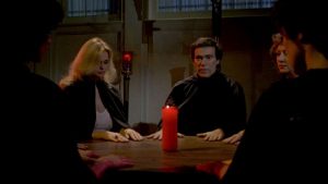 Un groupe d'hommes et de femmes vêtus tout de noir sont assis autour d'une table et d'une unique bougie rouge dans le film Cérémonie mortelle.