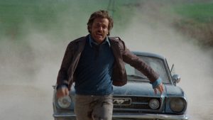Franco Nero court vers nous, poursuivi par une automobile à toute allure qui fait voler le sable dans les airs ; scène du film Un citoyen se rebelle.