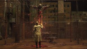 Une femme, vue de dos, fait face à un corps calciné, comme crucifié en pleine rue teintée de couleurs ocres, dans le film Silent Hill de Christophe Gans.