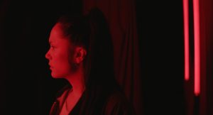 Plan rapproché-épaule sur une jeune femme brune, la mine très sérieuse et fermée, coiffée d'une queue de cheval, plongée dans une lumière rouge, dans une pièce indescriptible ; plan issu du film Bowling Saturne de Patricia Mazuy.