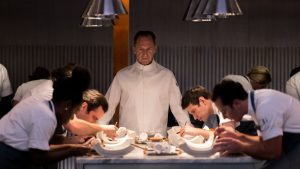 Le chef cuisinier interprété par Ralph Fiennes scrute ses apprentis cuistots, penchés sur leurs plats, alignés en deux rangées face à lui dans le film Le Menu.