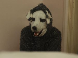 Plan rapproché-taille sur l'homme déguisé en chien du film Good Bye qui attend contre un mur blanc, la langue pendante.