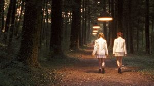 Deux jeunes filles vues de dos en uniforme d'écolière blanc marchent sur un sentir qui traverse une forêt ; étrangement des lampes éclairent le chemin, comme tombant du ciel ; plan issu du film Innocence de Lucile Hadzihalilovic.