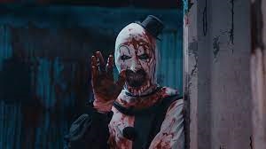 Plan rapproché-poitrine sur un Art le Clown souriant, faisant coucou, alors qu'il est abondamment tâché de sang, issu du film Terrifier 2.
