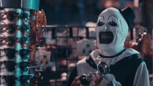 Art le Clown dans un magasin de farce et attrapes, à côté d'un étalage de lunettes de soleil ; il semble s'esclaffer ; scène du film Terrifier 2.