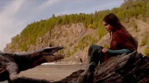 La jeune actrice Ariana Greenblatt est assise sur un rocher, au bord d'une plage, face à elle un dinosaure s'approche, sans animosité ; scène du film 65 : la Terre d'avant.